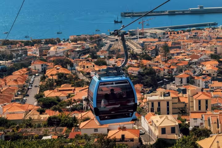 Um teleférico sobre os telhados alaranjados do Funchal, com o porto ao fundo.
