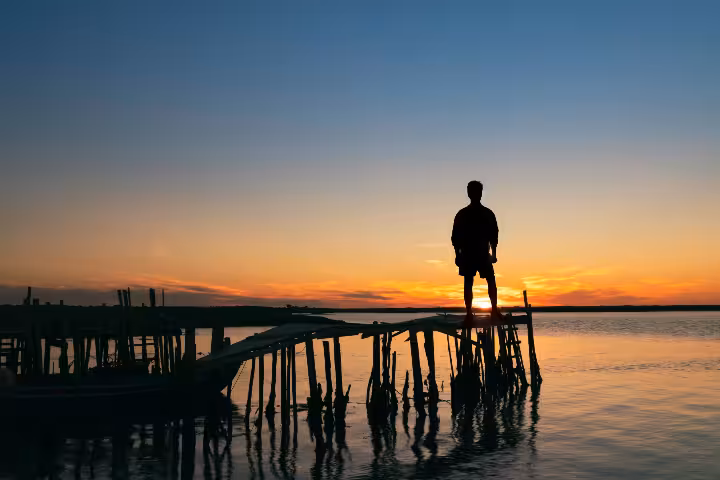 A lone traveler silhouetted against a breathtaking golden sunset over calm Portuguese waters, standing on a rustic wooden pier for a soul-stirring Portugal travel experience with ToursXplorer.