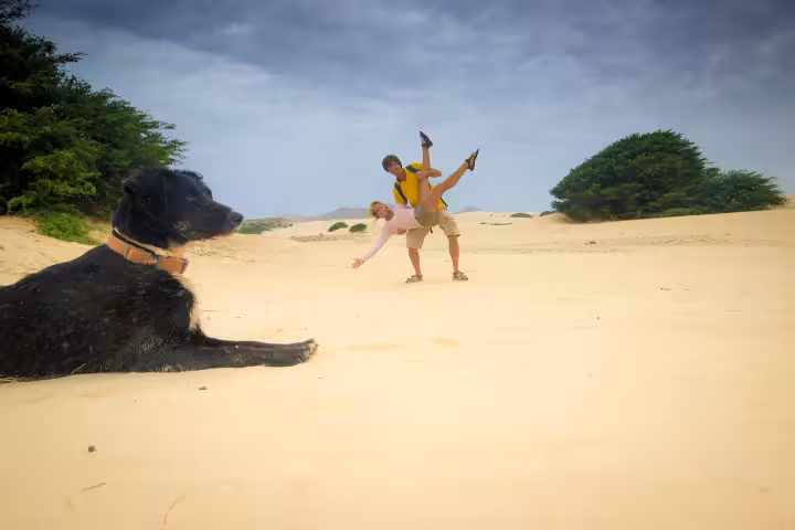 A playful couple enjoying an adventurous acrobatic moment on the golden sand dunes of Cape Verde with a black dog in the foreground, surrounded by lush green trees and a clear blue sky, capturing the spirit of an adventure holiday in Cape Verde.