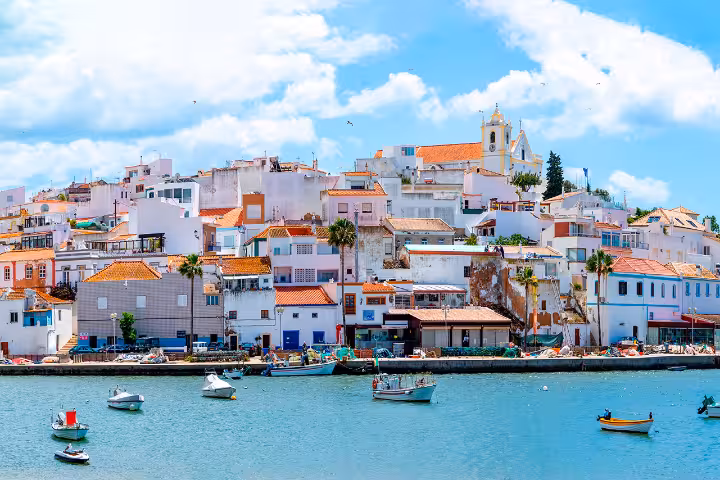 Panoramic waterfront view of Ferragudo village in Algarve Portugal featuring traditional whitewashed houses with terracotta roofs a hilltop church and fishing boats along the scenic harbor perfect for tours and travel exploration.