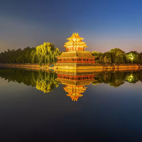 Vista panorâmica da torre de canto iluminada e do fosso da Cidade Proibida ao entardecer em Pequim, China, exibindo a arquitetura histórica e os reflexos serenos.