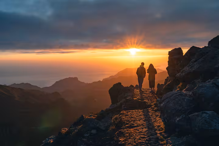 Two hikers standing on the rocky summit of Pico Ruivo at golden sunset, enjoying breathtaking panoramic views of Madeira's misty mountain peaks along the scenic Pico do Areeiro to Pico Ruivo hiking trail.