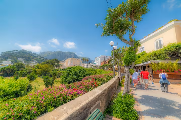 Tourists strolling along the vibrant flower-lined promenade of Capri Island with lush gardens, elegant buildings, and dramatic mountain backdrop on a sunny summer day during an Italian Riviera and Amalfi Coast tour.