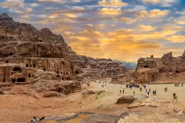 Panoramic daytime view of Petra Jordan's ancient Nabataean rock-carved tombs and temples with tourists exploring the UNESCO World Heritage Site under a dramatic golden sunset sky, perfect for guided Petra day tours and sightseeing adventures.