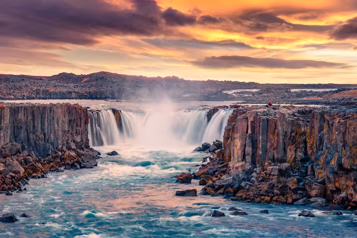 A breathtaking panoramic view of Selfoss Waterfall in Iceland during the midnight sun, with golden summer skies illuminating the dramatic basalt canyon and powerful cascading water, perfect for Iceland summer vs winter travel planning.