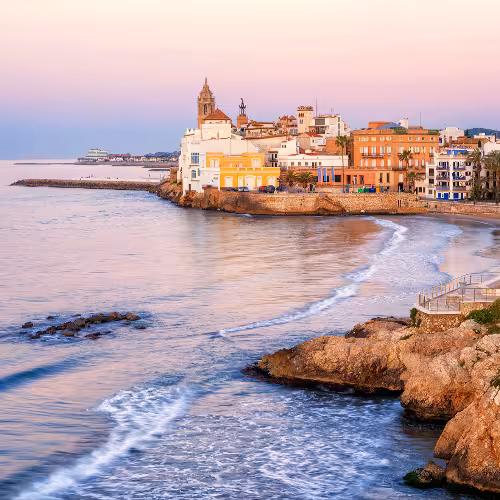 Vista do pôr do sol na cidade velha de Sitges e no calçadão à beira-mar, na Catalunha, Espanha, com edifícios coloridos à beira-mar, palmeiras e praia de areia — uma das melhores coisas para fazer perto de Barcelona.