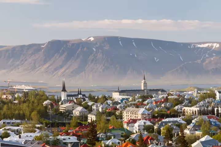 Panoramic aerial view of Reykjavík city skyline with colorful rooftops, historic church spires, lush green trees, and snow-capped Esja mountain in the background, perfect for a private driving tour or walking tour in Iceland.