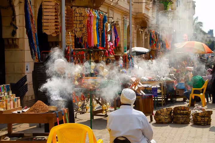 A local vendor in a traditional white turban surrounded by aromatic incense smoke, golden lanterns, and vibrant spice stalls at Khan el-Khalili bazaar in Cairo, Egypt, a must-visit cultural sightseeing experience on guided Cairo tours.