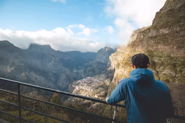 A solo traveler in a blue jacket gazing at the breathtaking misty mountain valleys and a secluded village below from a scenic viewpoint in Madeira, perfect for solo travel adventures and self-discovery tours.