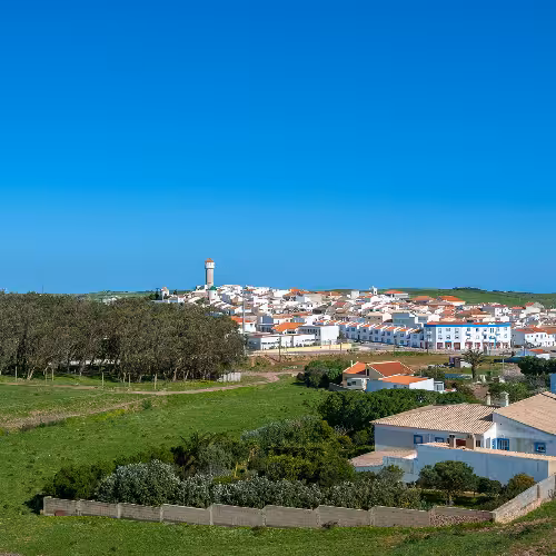 A picturesque view of Vila do Bispo in the Algarve region of Portugal, showcasing traditional whitewashed houses with red roofs surrounded by lush greenery under a clear blue sky.
