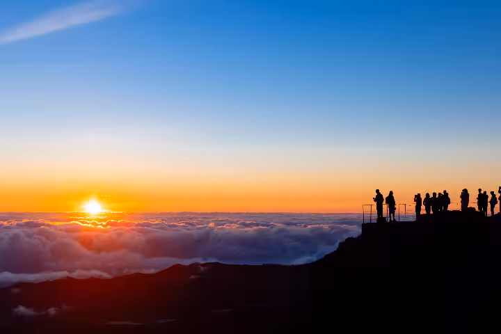 Silhouettes of tourists gathered at the Pico do Areeiro summit viewpoint in Madeira, witnessing a breathtaking sunrise above a sea of clouds with vibrant orange and golden sky hues.