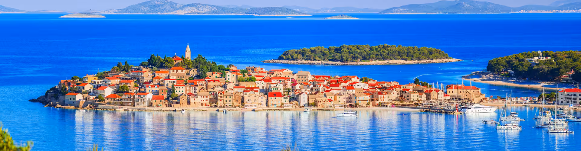 Panoramic view of a historic coastal town with red-roofed stone houses and marina yachts on the Adriatic Sea in Dalmatia, Croatia, surrounded by turquoise bays and nearby islands.