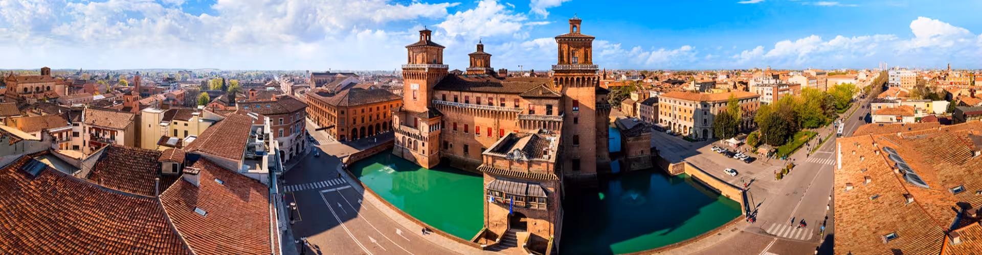 Panoramic view of Castello Estense and the historic center of Ferrara, Italy, with medieval towers, canals and terracotta rooftops under a bright blue sky