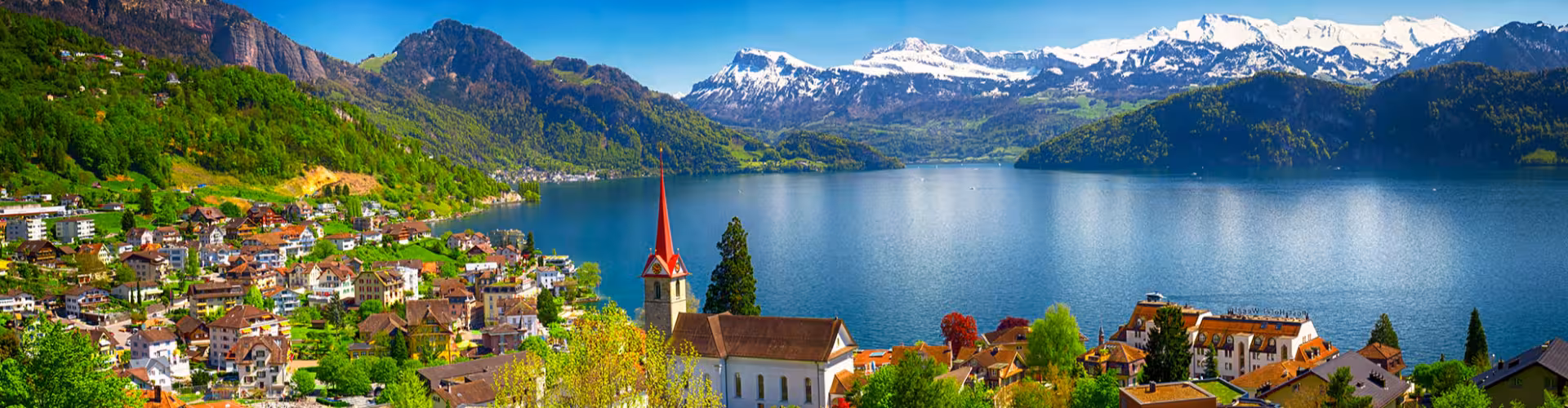 Panoramic view of Lake Lucerne, Switzerland with Lucerne old town rooftops, a lakeside church spire, and snow-capped Swiss Alps reflecting in the water.