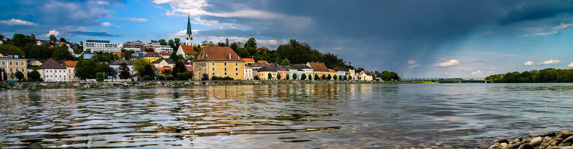 Danube River waterfront panorama in Mauthausen, Upper Austria, with colorful historic townhouses and church spire under dramatic skies near Linz.