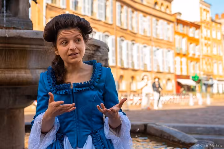 Guide in blue 18th century costume gesturing near a fountain in Toulouse Saint-Étienne district