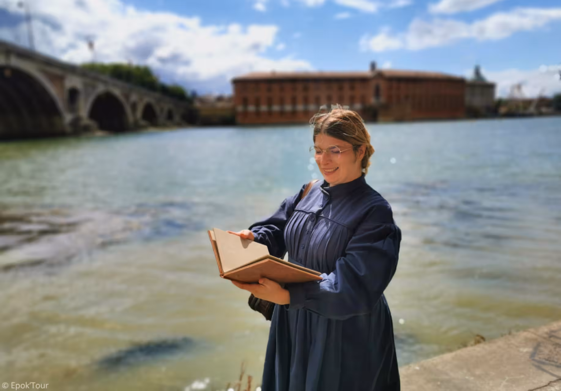 Guide in 19th-century costume reading a book by the Garonne River in Toulouse with a stone bridge behind