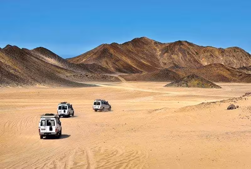 Three white 4x4 jeeps driving across sandy desert with rocky mountains in Marsa Alam, Egypt