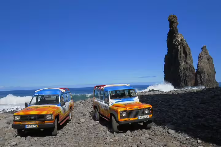 Two colorful 4x4 vehicles parked on rocky terrain near dramatic sea stacks on a sunny day during the West volcanic pools tour.