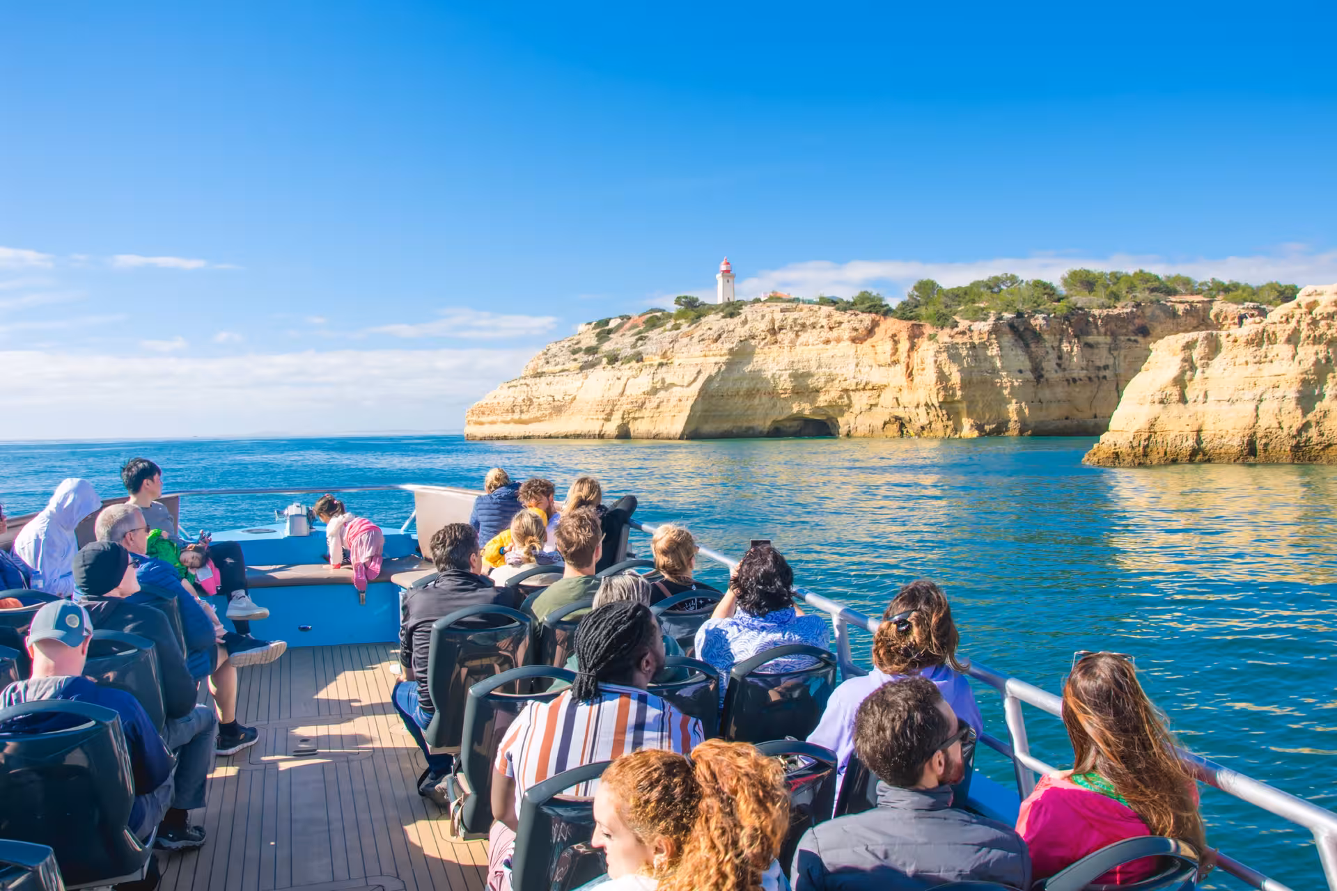 Visitors on a boat tour admire the scenic Algarve coastline featuring cliffs and a lighthouse under a bright blue sky.