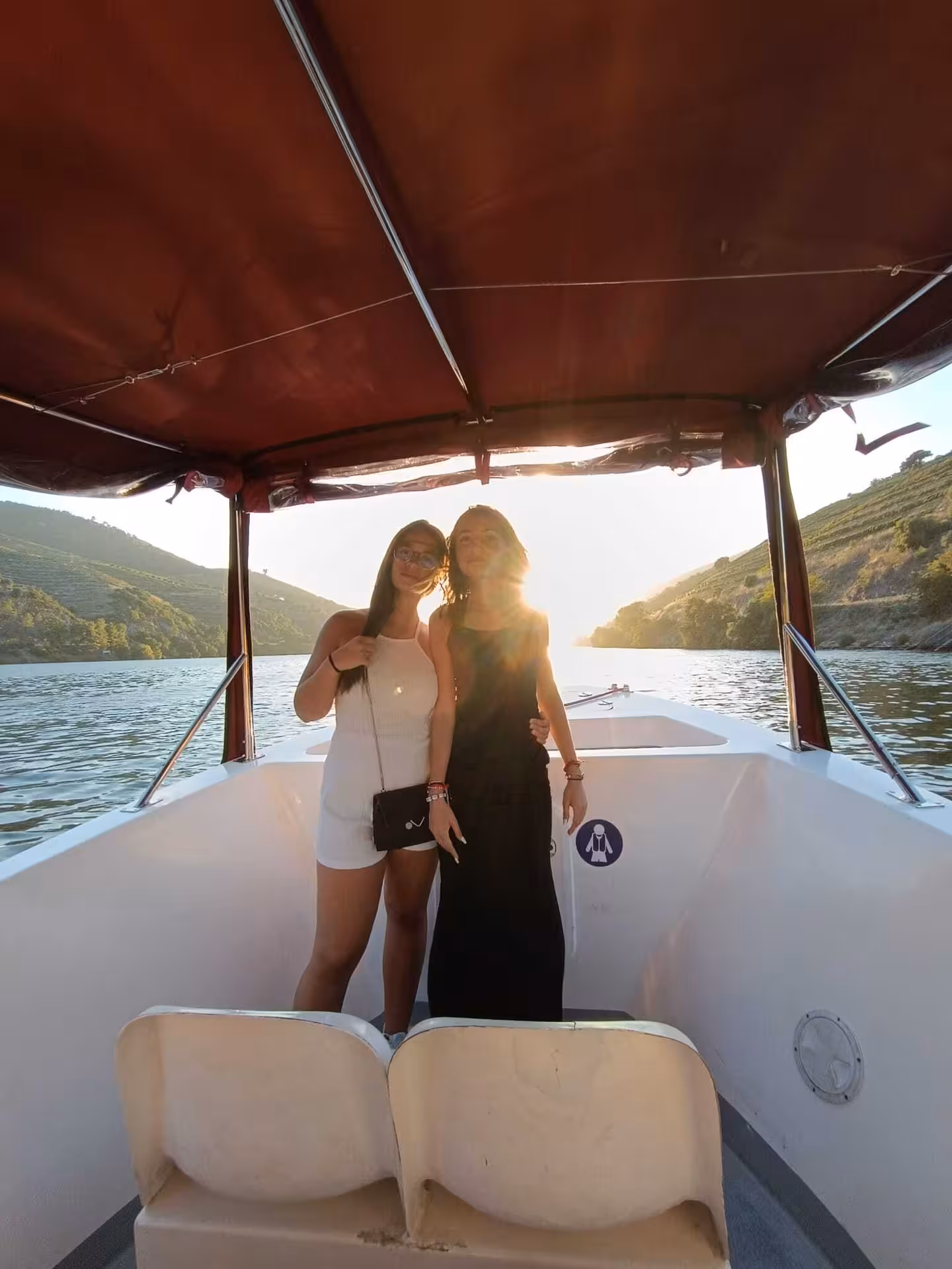 Two women enjoying a scenic Douro River sunset boat tour from Pinhão with golden vineyard hills and shimmering river waters in the background