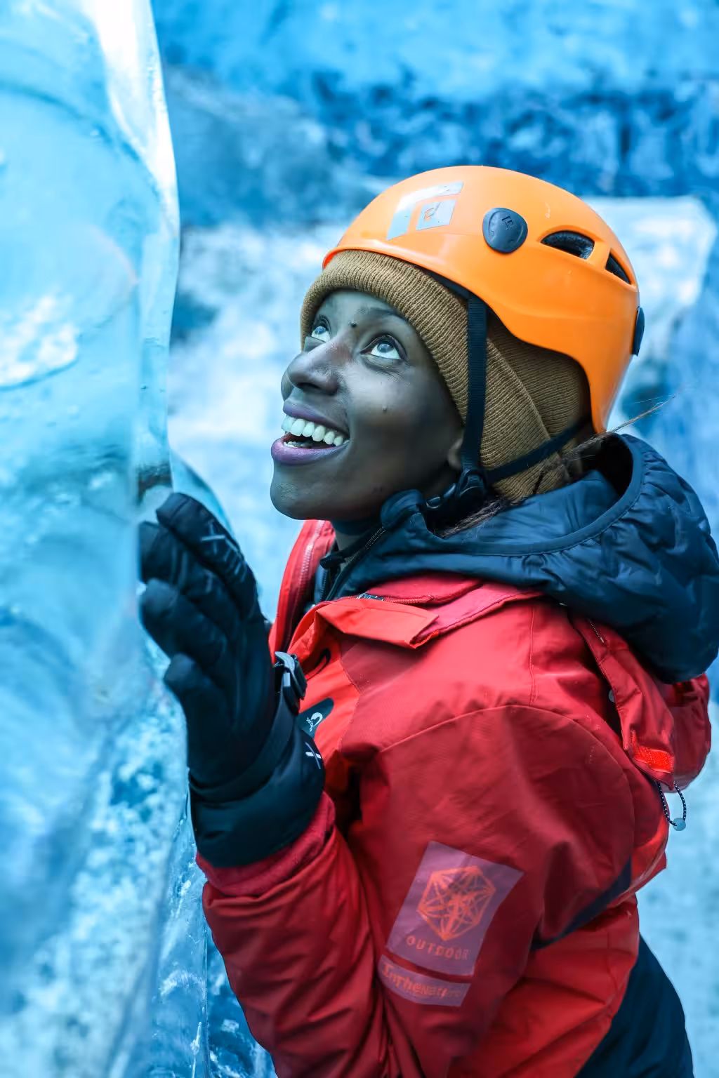 Smiling woman in red jacket and orange helmet touching blue glacier ice wall on Vatnajokull ice cave guided tour