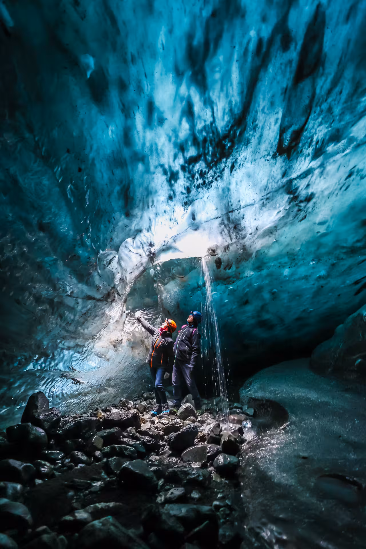 Two guided tour guests wearing helmets marvel at a waterfall inside the vivid blue Vatnajokull ice cave Iceland