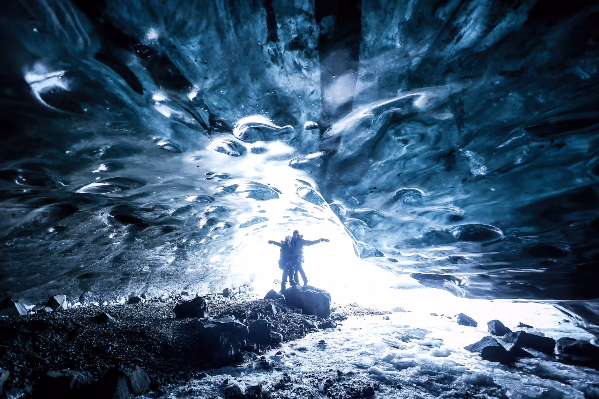 Two travelers pose joyfully inside a stunning Vatnajokull ice cave with glowing blue glacial walls on a guided Iceland tour.