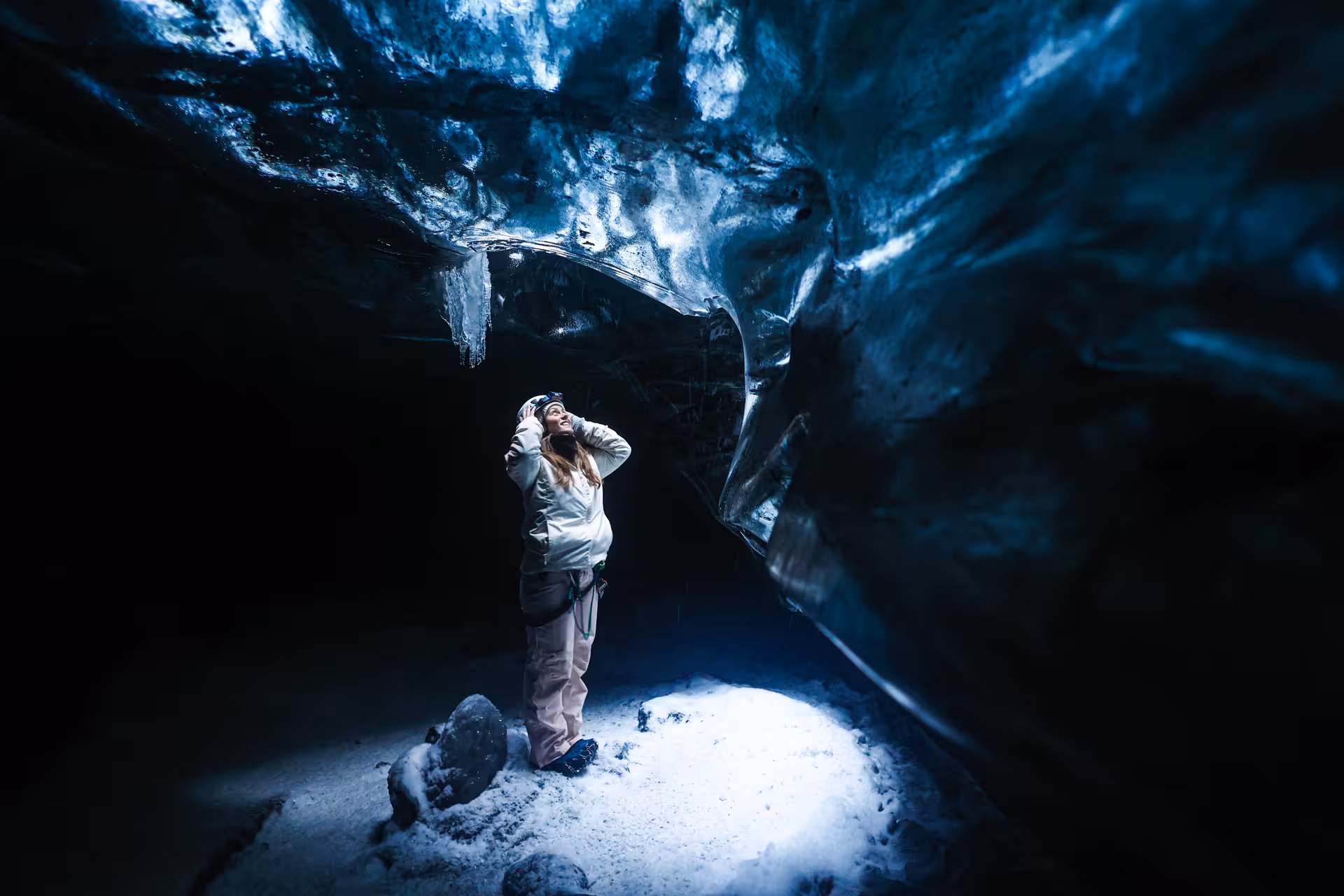 Woman in white winter gear marveling at the dark blue glacial walls inside a Vatnajokull ice cave on a guided Iceland tour.