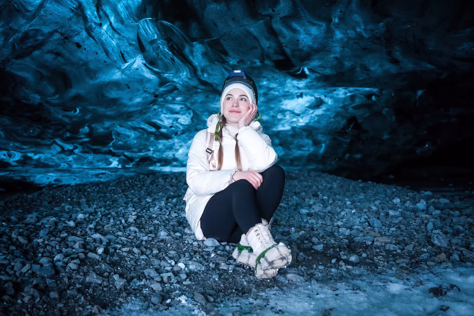 Young woman in white jacket sits on rocky ground admiring glowing blue ice walls of Vatnajokull ice cave Iceland