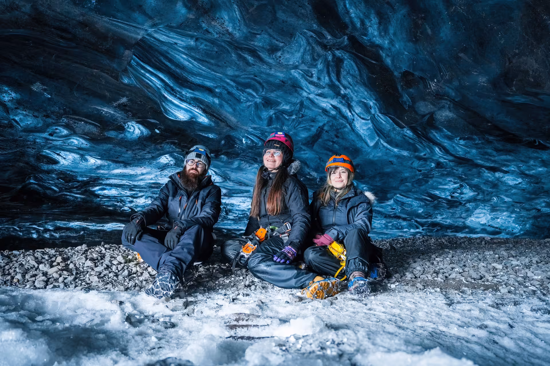 Three tourists in helmets and gear sit inside a stunning blue Vatnajokull ice cave on a guided Iceland tour