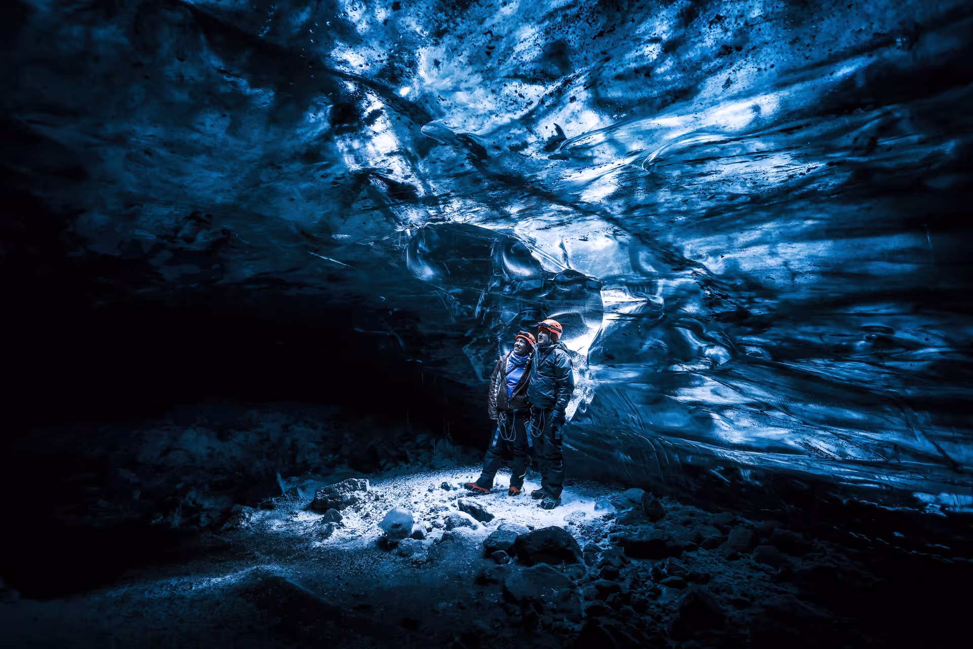 Two helmeted adventurers gaze up at glowing blue ice formations inside a Vatnajokull ice cave guided tour Iceland