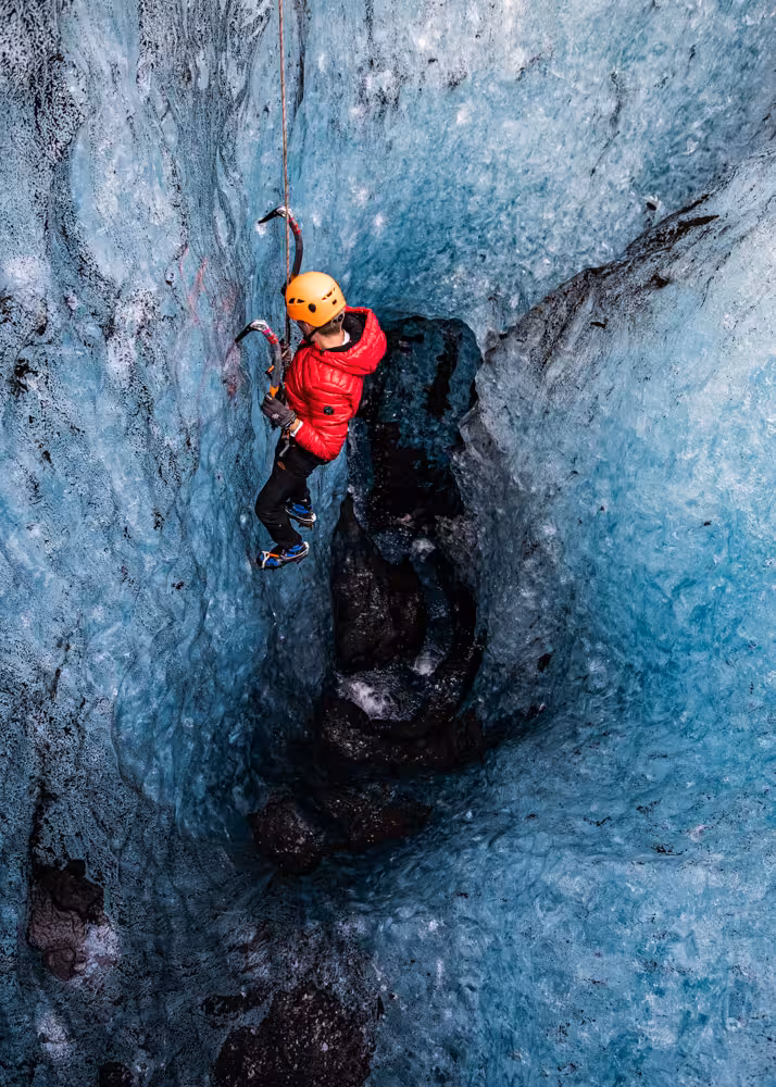 Adventurer in red jacket and orange helmet ascending a dramatic blue glacial ice crevasse using ice axes and crampons