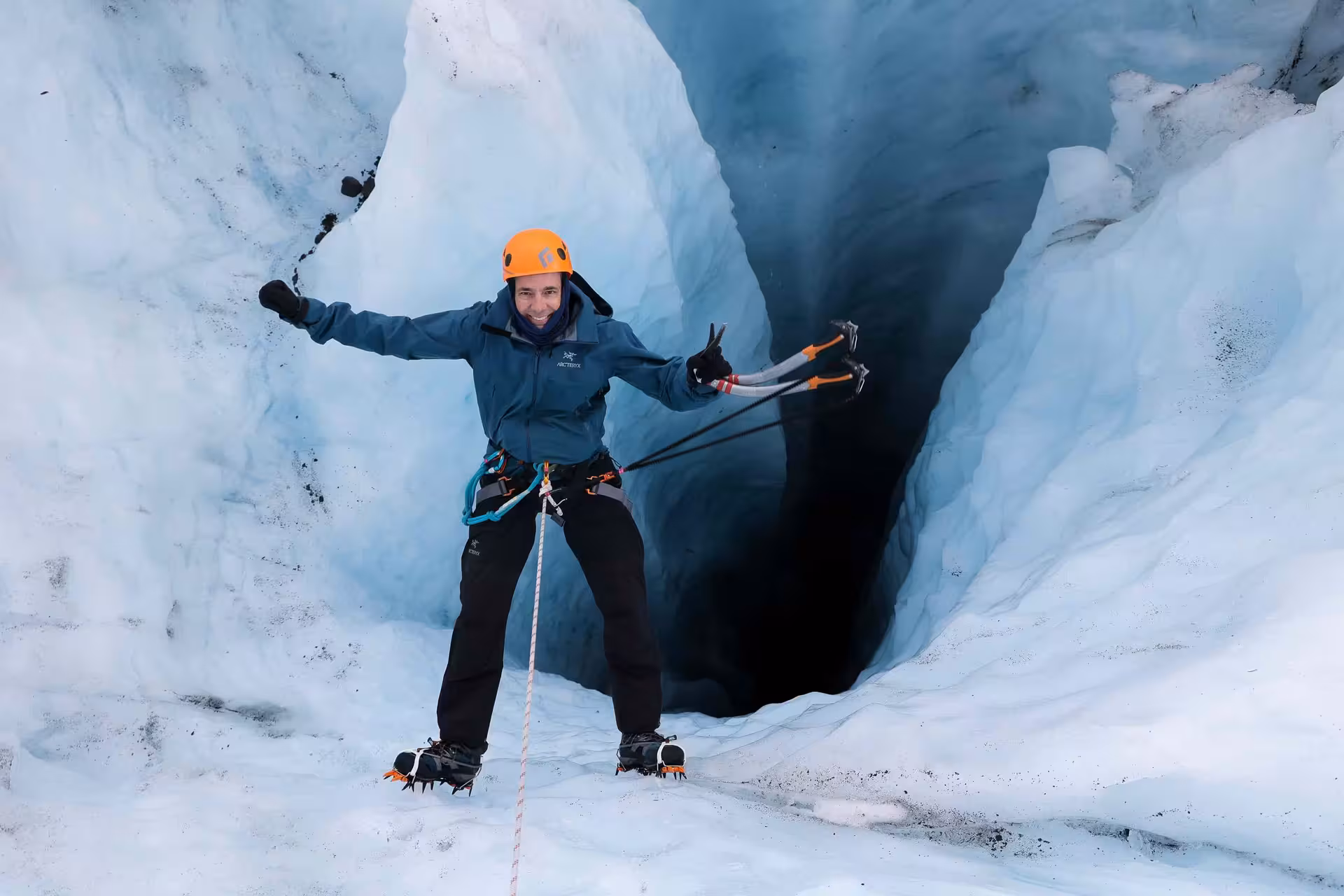 An excited adventurer in full gear poses with an ice axe at the entrance of a glacier cave on a guided ice climbing tour.