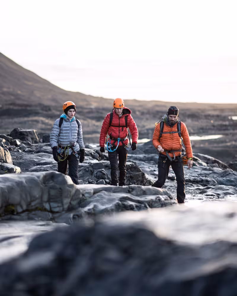 Three glacier ice climbing tour participants in helmets and harnesses trekking across rocky terrain toward the glacier.