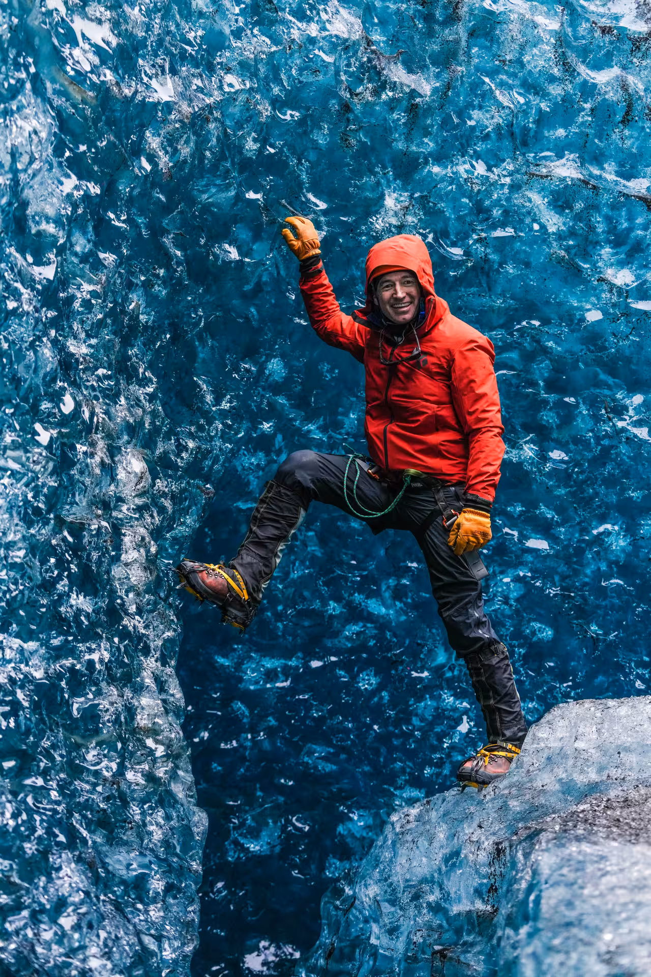 Smiling climber in red jacket scaling a vivid blue glacial ice wall with crampons and ice axe on a guided photo tour.