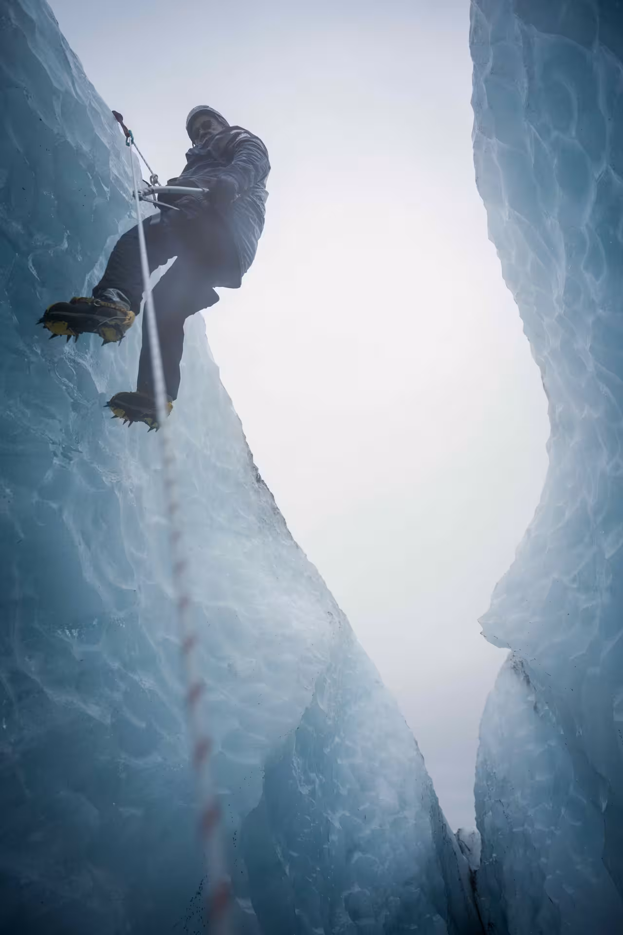 Low-angle view of a glacier ice climber with crampons and rope ascending a narrow blue ice crevasse on a guided tour.