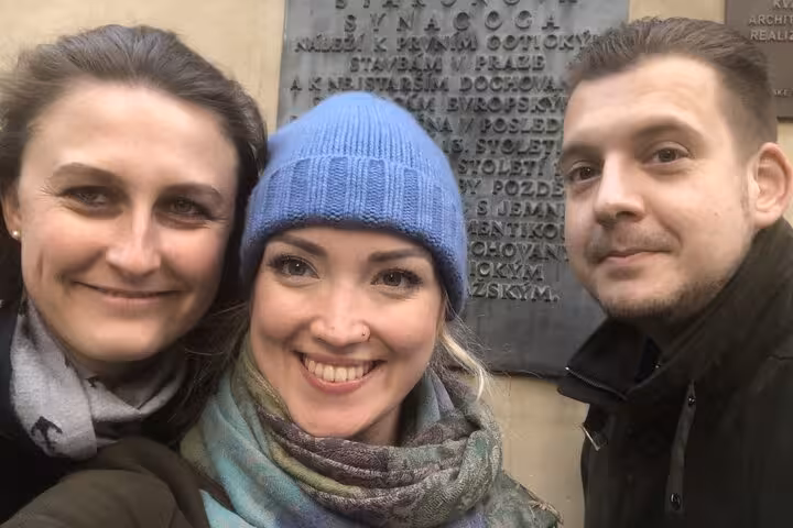 Three friends smiling in front of historic Prague synagogue plaque during an outdoor escape game walking tour