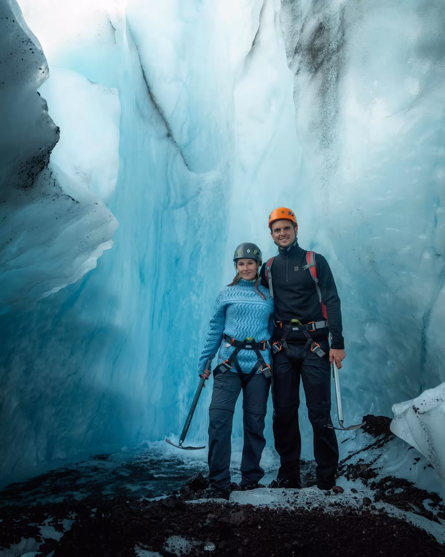A couple in helmets and harnesses poses inside a breathtaking blue ice cave during a Falljokull Glacier hike in Skaftafell Iceland
