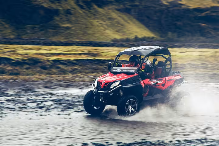 Driver in orange gear waves from a Can-Am buggy on rocky terrain in Iceland with green hills behind