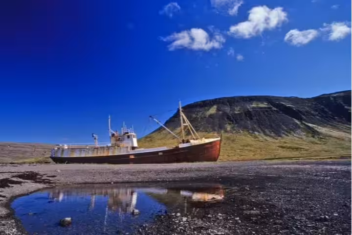Abandoned rusty shipwreck beached on Iceland's rocky shore with dramatic mountain backdrop and blue sky