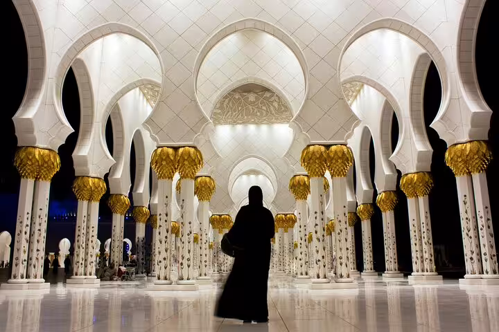 Interior of a majestic mosque in Abu Dhabi with intricate arches, golden columns, and a silhouetted visitor.