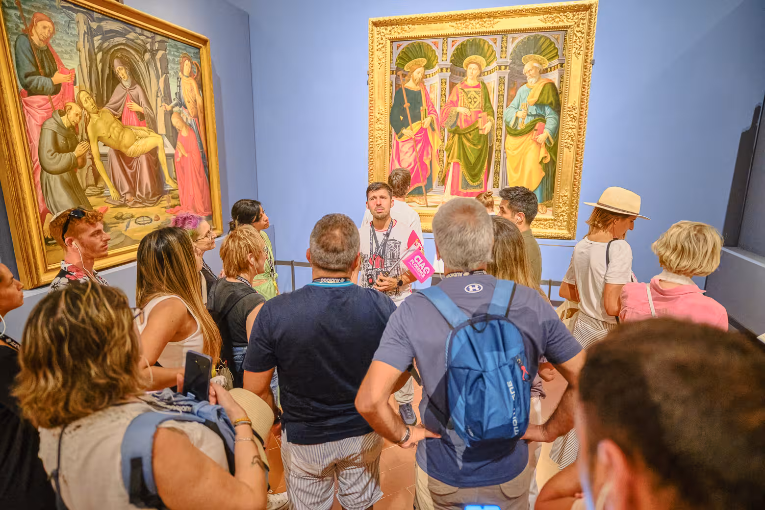 Visitors gathered around a guide in Accademia Gallery, Florence, admiring detailed Renaissance paintings.