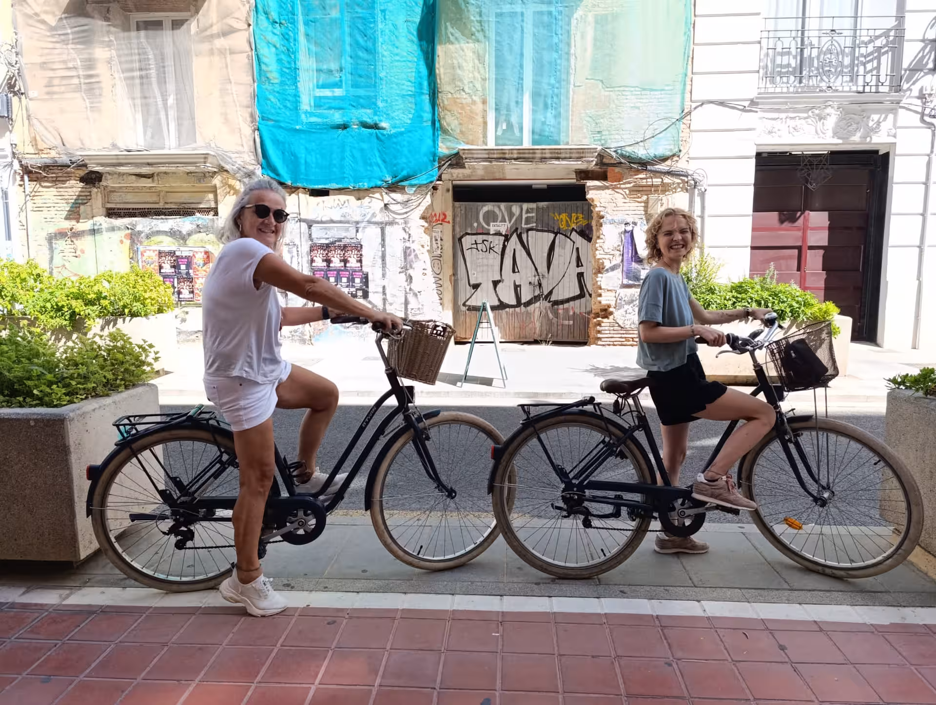 Two adults on comfortable city bike rentals with baskets, stopped on a lively street for easy sightseeing rides