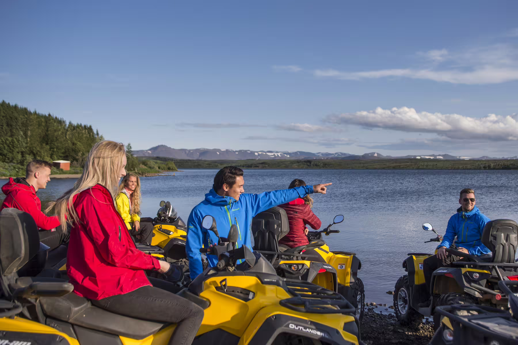 Group of adventurers on yellow ATVs by a scenic lake with mountains in the background, enjoying an ATV and caving tour.
