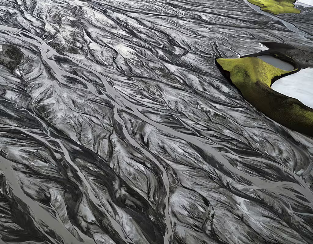Aerial view of braided glacial river channels with dark volcanic sediment and green moss island in South Iceland