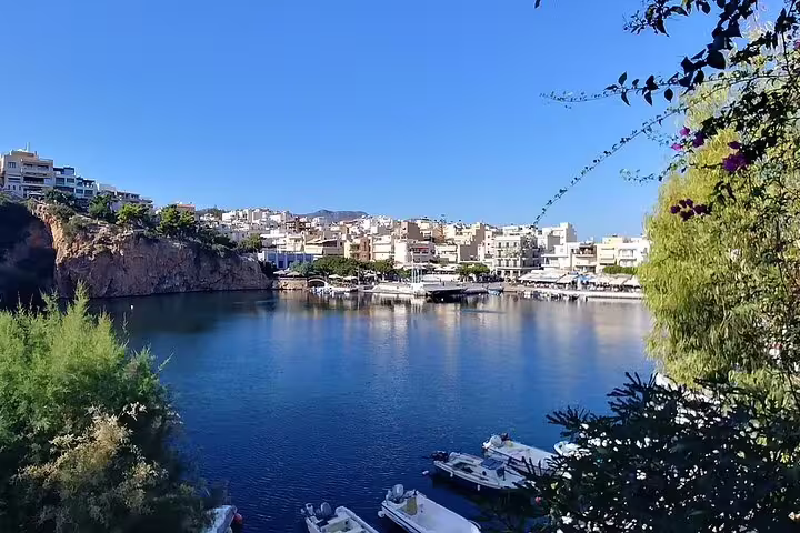 Scenic view of Agios Nikolaos harbor with boats, surrounded by lush greenery and charming buildings under a clear blue sky.