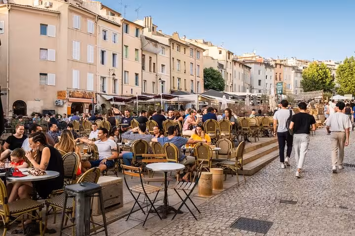 Crowded outdoor café terrace with wicker chairs in a sunny Aix-en-Provence square lined with pastel buildings