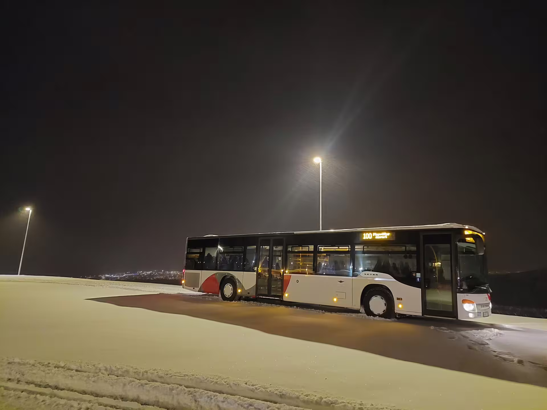 Setra bus route 100 parked in snowy lot at night, illuminated by streetlights