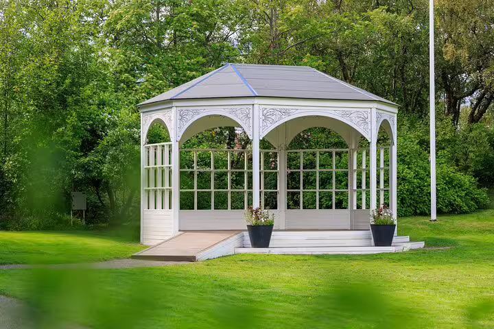 White ornate gazebo with arched windows surrounded by lush green trees in Akureyri Botanical Gardens